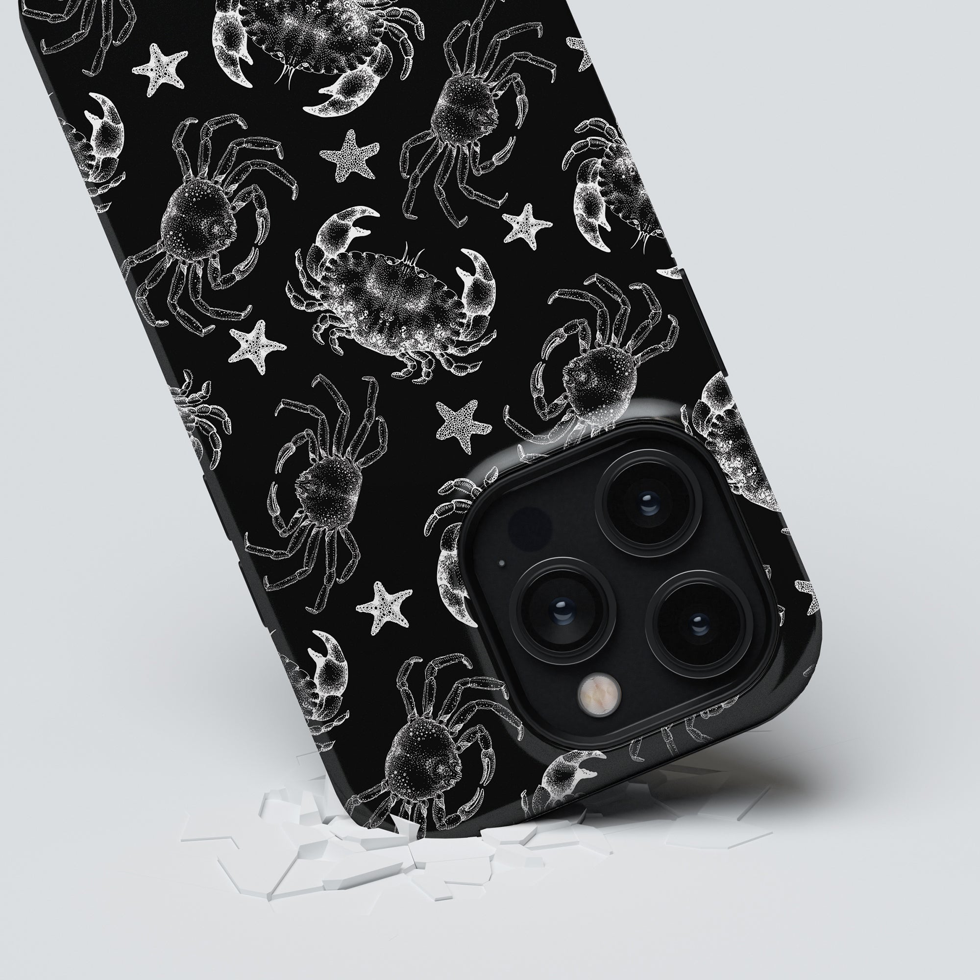 A smartphone from the Ocean Collection with a robust Black Crab - Tough Case featuring a white crab and starfish pattern is lying on a white surface. The Black Crab - Tough Case prominently displays the phone's camera lenses.