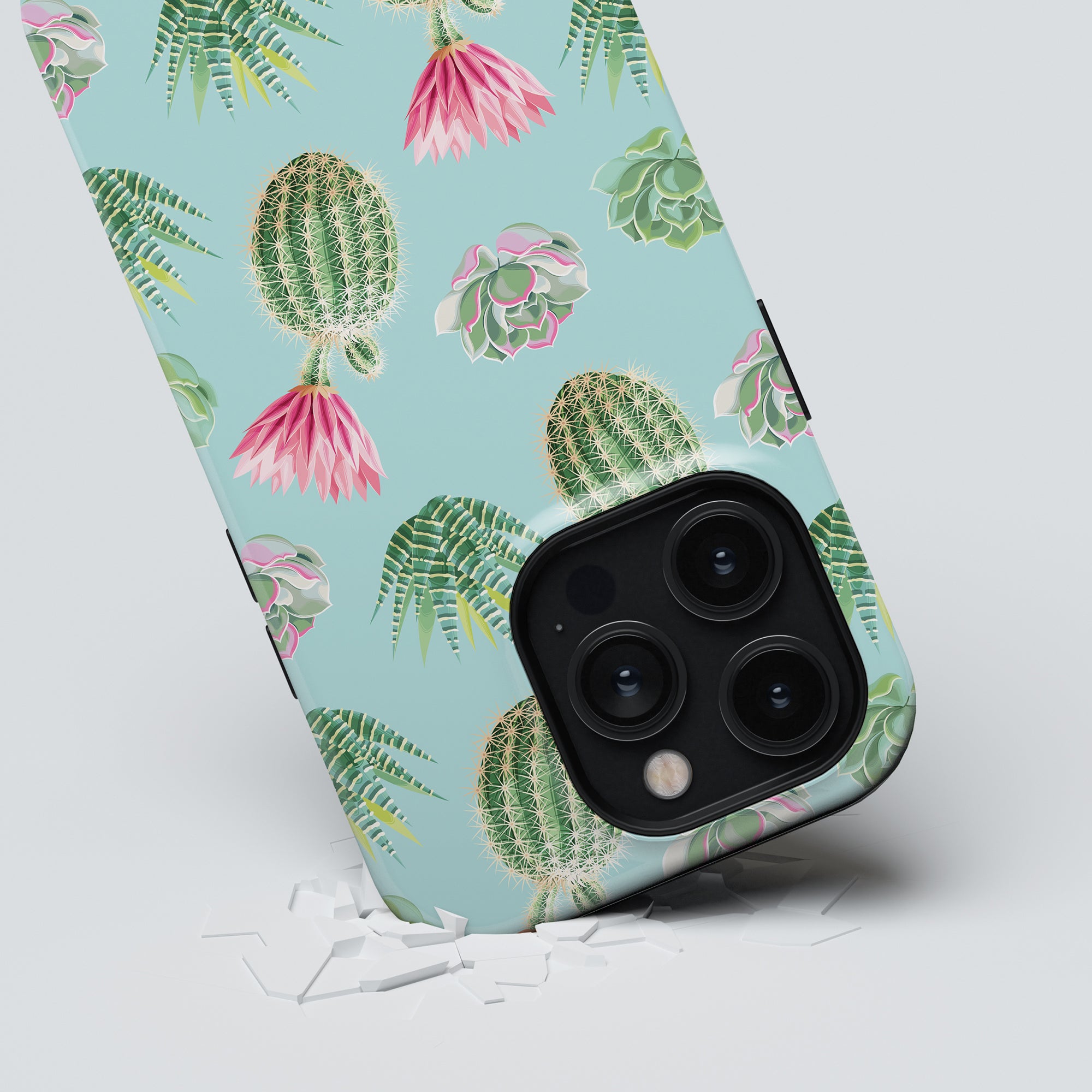 A "Cactus Splash - Tough Case" from the Exotic Fruits Collection, adorned with a tropical cactus design, rests on a white surface scattered with small fragments. The tough case phone prominently features three camera lenses.