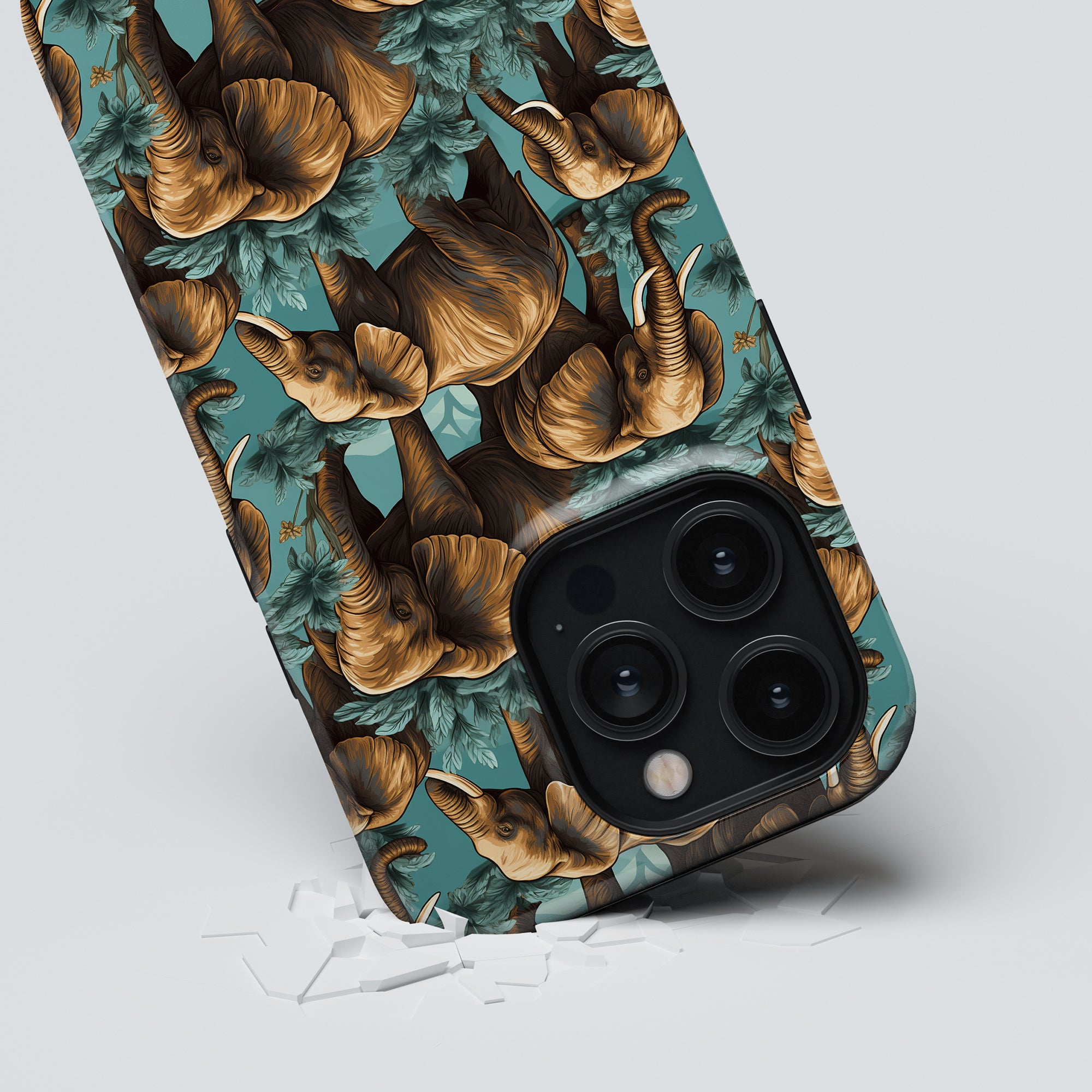 A smartphone with an intricate elephant design from the Hathi - Tough Case is placed on a light surface, surrounded by scattered pieces of shattered glass.