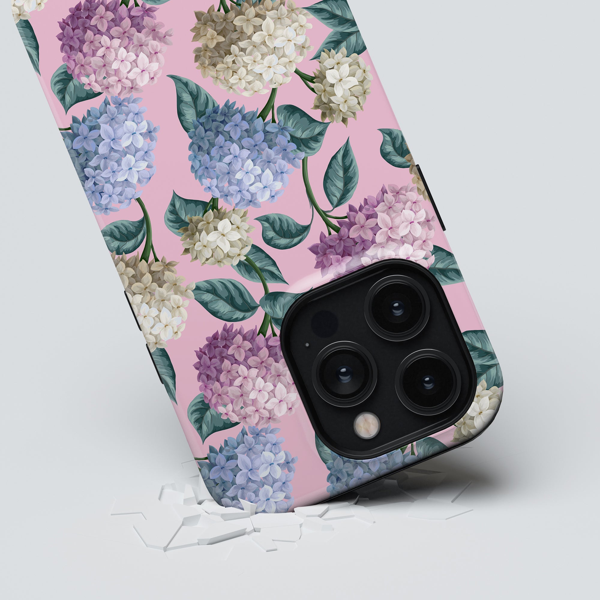 A smartphone with a Hydrangea - Tough Case from the Summer Collection is placed on a white surface. The phone's camera is prominently visible, showcasing three lenses. Small white fragments are scattered near the phone, highlighting its robust smartphone protection.