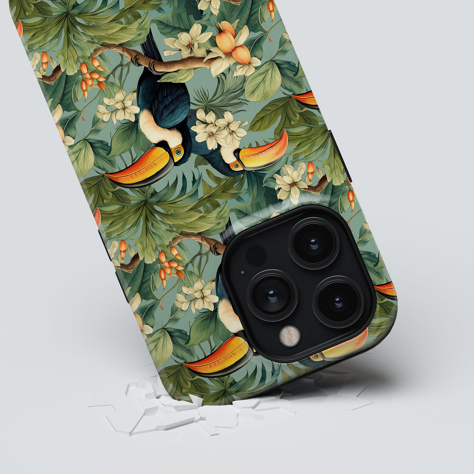 The Ramphastos - Tough Case, featuring a tropisk elegans theme with vibrant tukaner and lush leaves, rests at an angle on a light background, elegantly surrounded by a small scatter of white pieces beneath it.