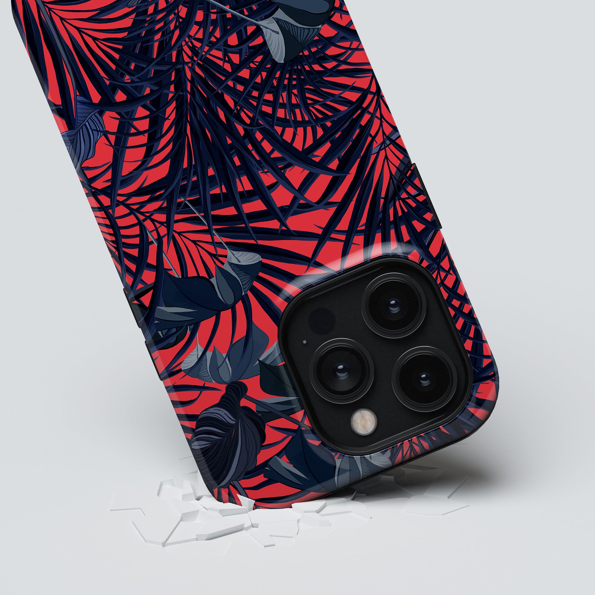 A smartphone with a Red Tropics - Tough Case featuring red and dark palm leaves is propped up on a white surface, with a visible crack underneath its lower right corner.
