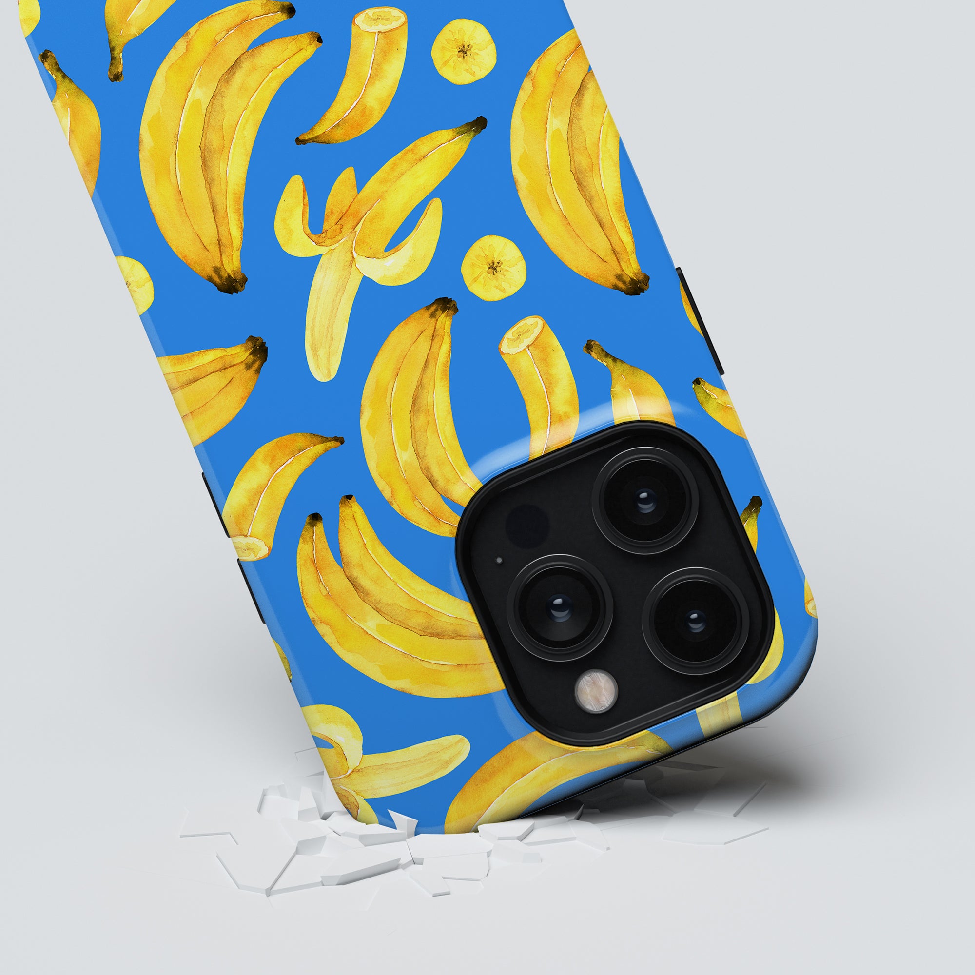 A phone encased in the "Ripe Bananas - Tough case" from the Exotic Fruits Collection, characterized by a blue backdrop adorned with ripe banana designs, rests on a white surface scattered with small debris at its base.