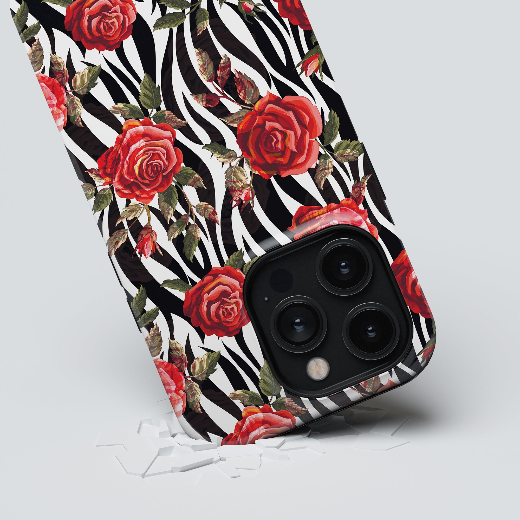 A smartphone encased in a Rose - Tough case from the Floral Collection, featuring a zebra and red roses pattern, lies on its corner on a light surface with small white fragments scattered around it.