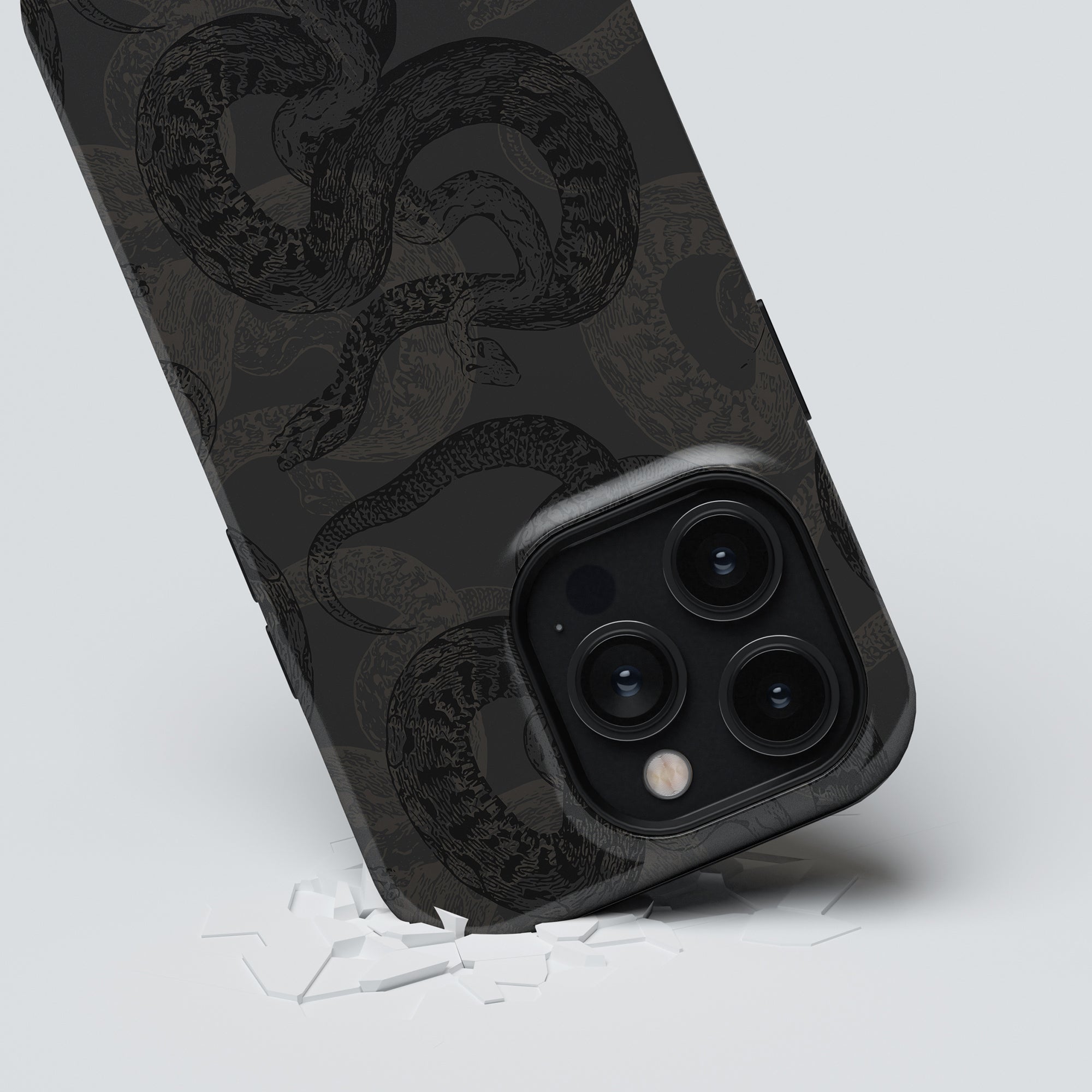 A smartphone encased in the Serpent - Tough case from the Snakeskin Collection is propped on a surface, showcasing a close-up view of its triple-camera system. Small white shards are scattered around its base.