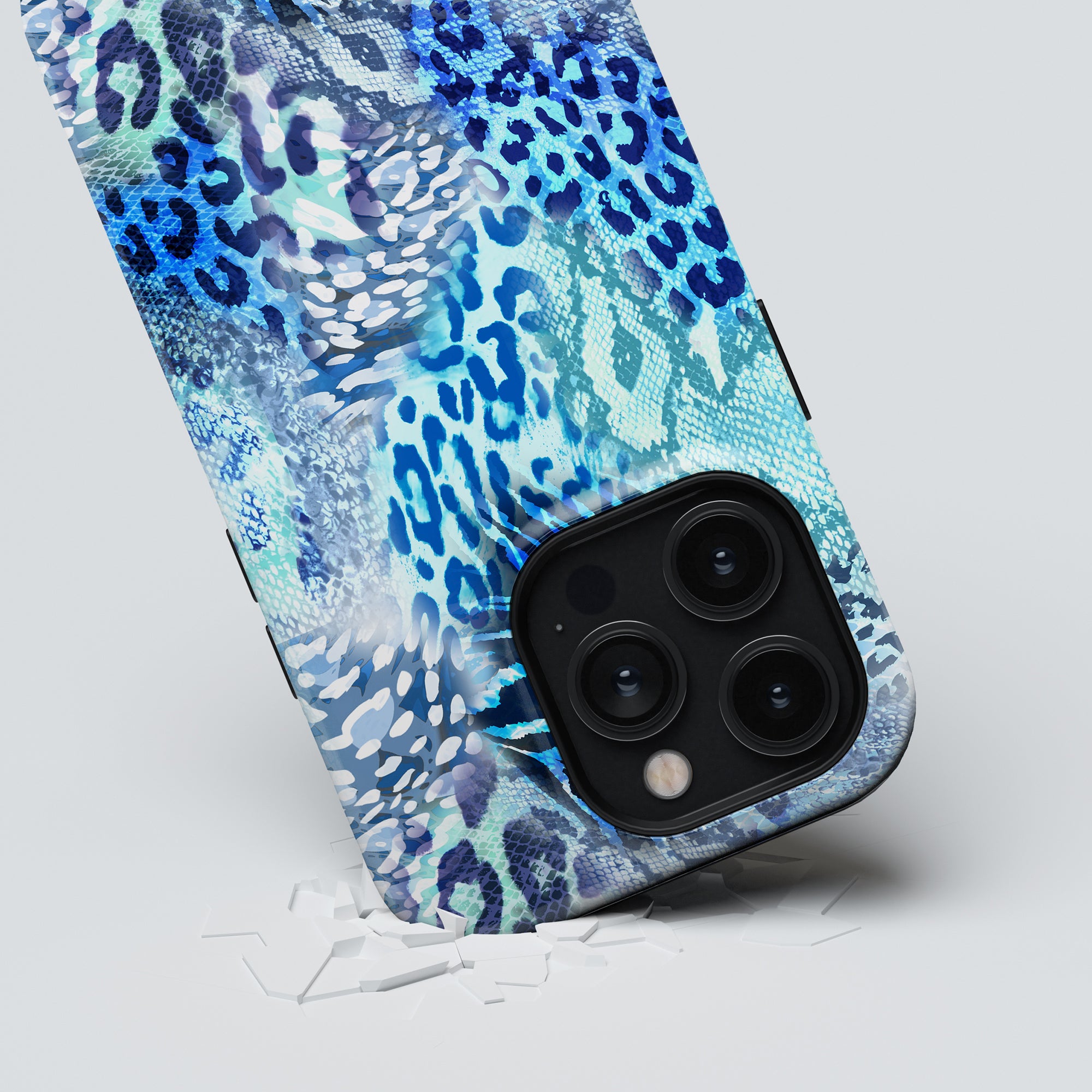 A smartphone with a Snow Leopard - Tough Case featuring a blue and white animal print rests against a white surface, surrounded by broken pieces of glass, indicating the phone has been dropped.