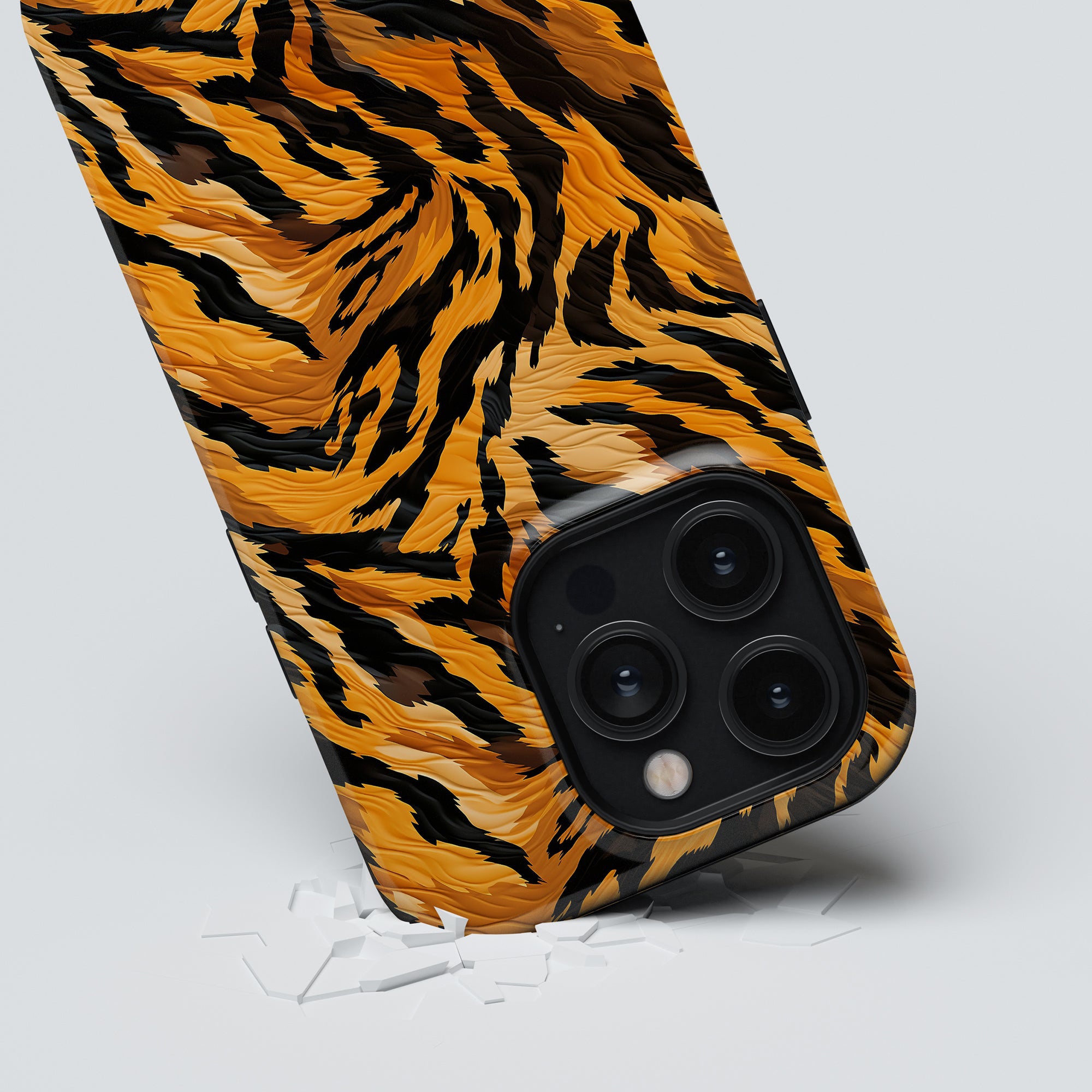 A smartphone with a Tiger Fury - Tough case featuring a striking tiger stripe pattern is shown leaning on a white surface. The camera lenses are visible, and there are small broken pieces on the surface near the phone, indicating its need for optimal protection.