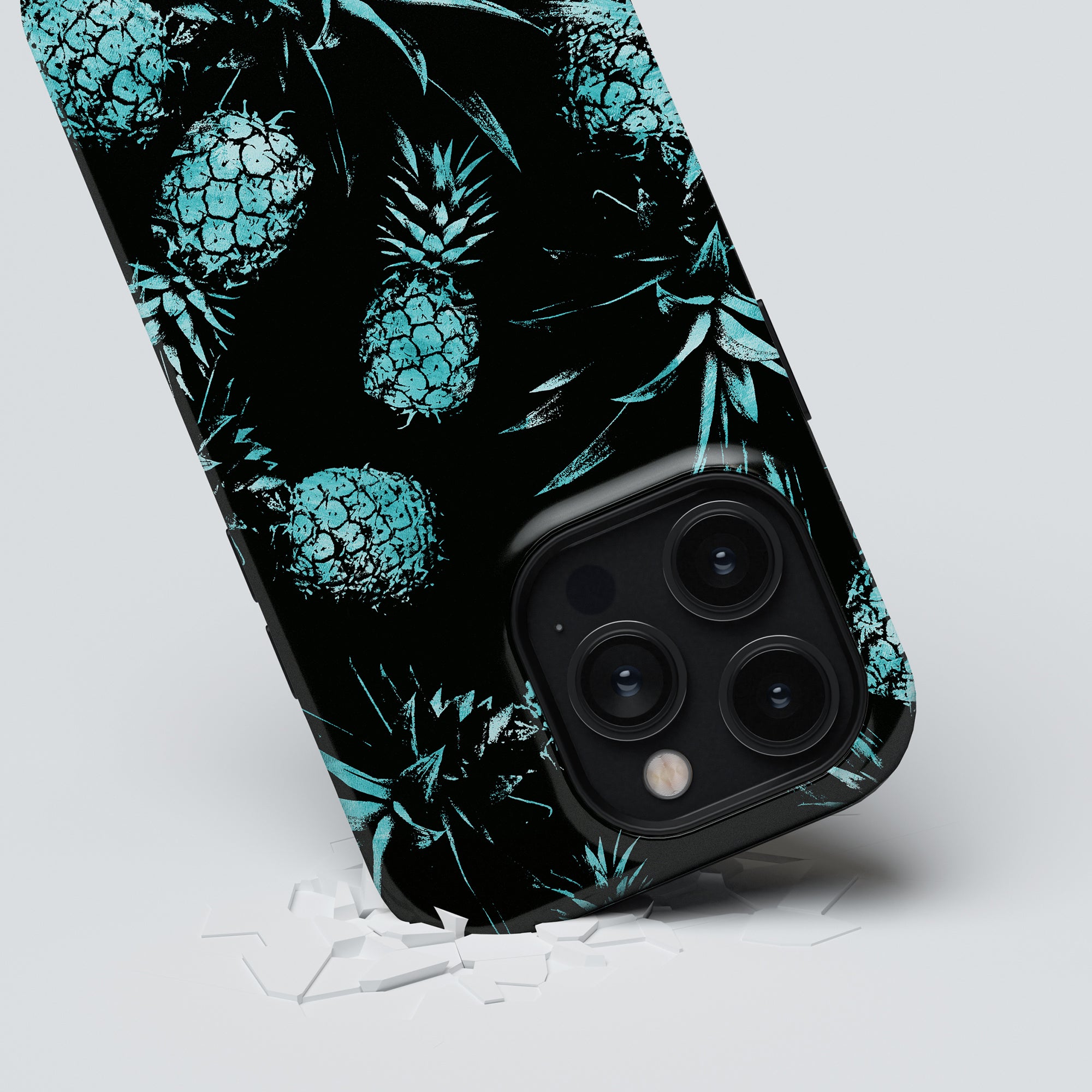 A smartphone with a Turquoise Pineapples - Tough Case from the Exotic Collection lies on a white surface with scattered shards of glass around it.