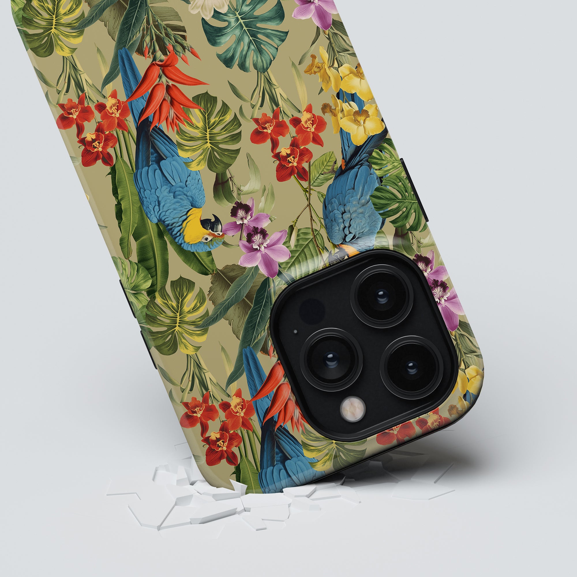 A smartphone with a Verdant Beauty - Tough Case from the Birds Collection, displaying vibrant parrots and flowers, is placed on a light surface with some white bits nearby. The phone's three rear camera lenses are visible.